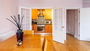 Una zona de comedor con una mesa, sillas, un florero con ramas negras y una cocina al fondo.