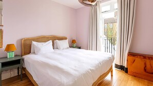 Un dormitorio con una cama grande, dos mesitas de noche con lámparas, un cabecero de madera y vistas al exterior a través de una ventana con cortinas.