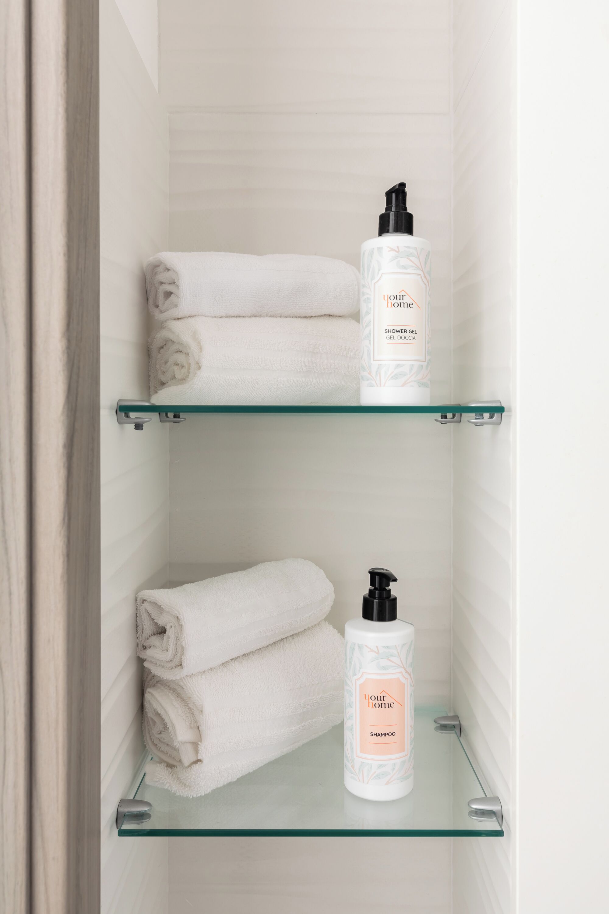 A bathroom with two glass shelves, each holding a rolled white towel and a bottle of shower gel and shampoo.