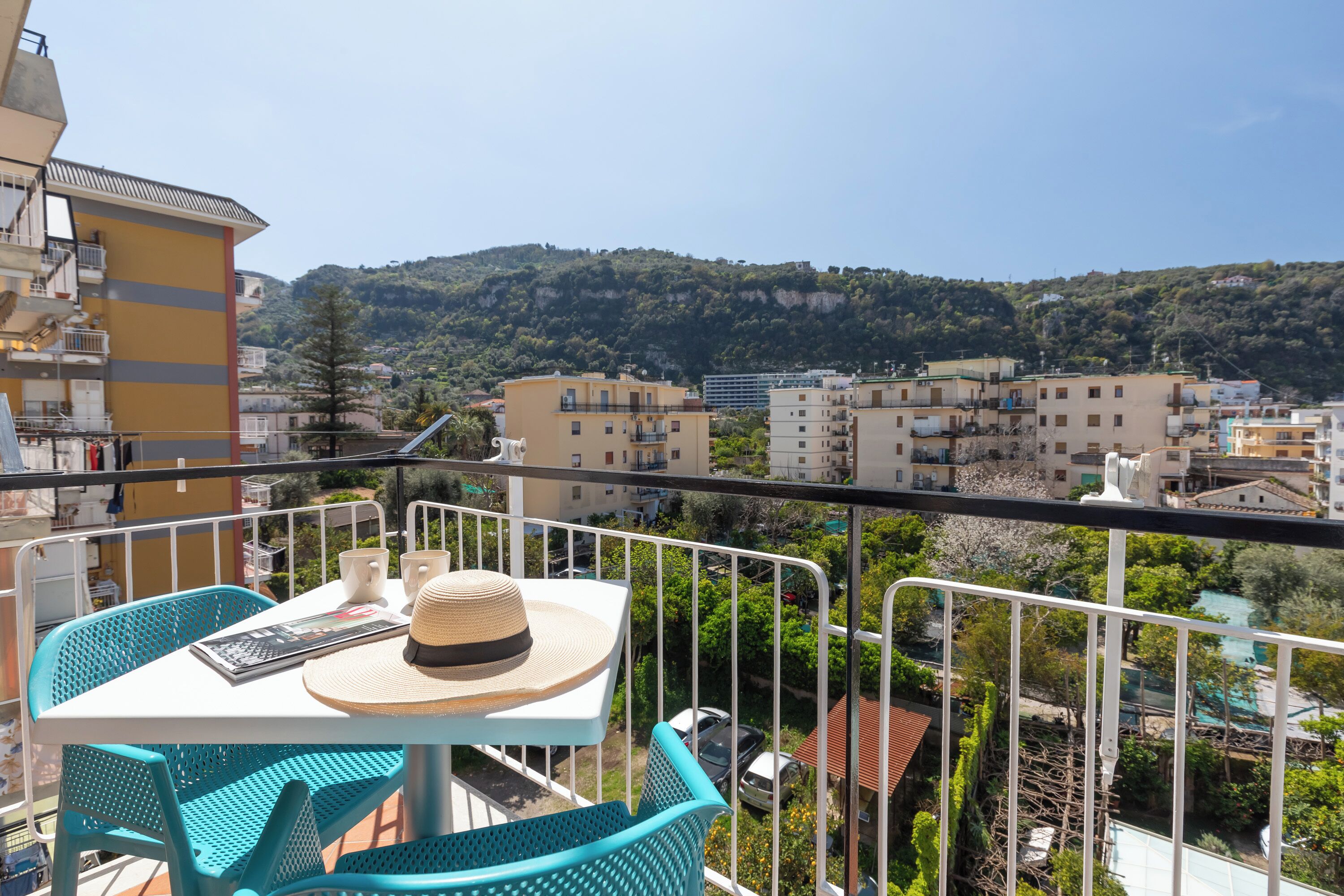 A balcony with turquoise chairs and a table, overlooking a cityscape with buildings and a hill.