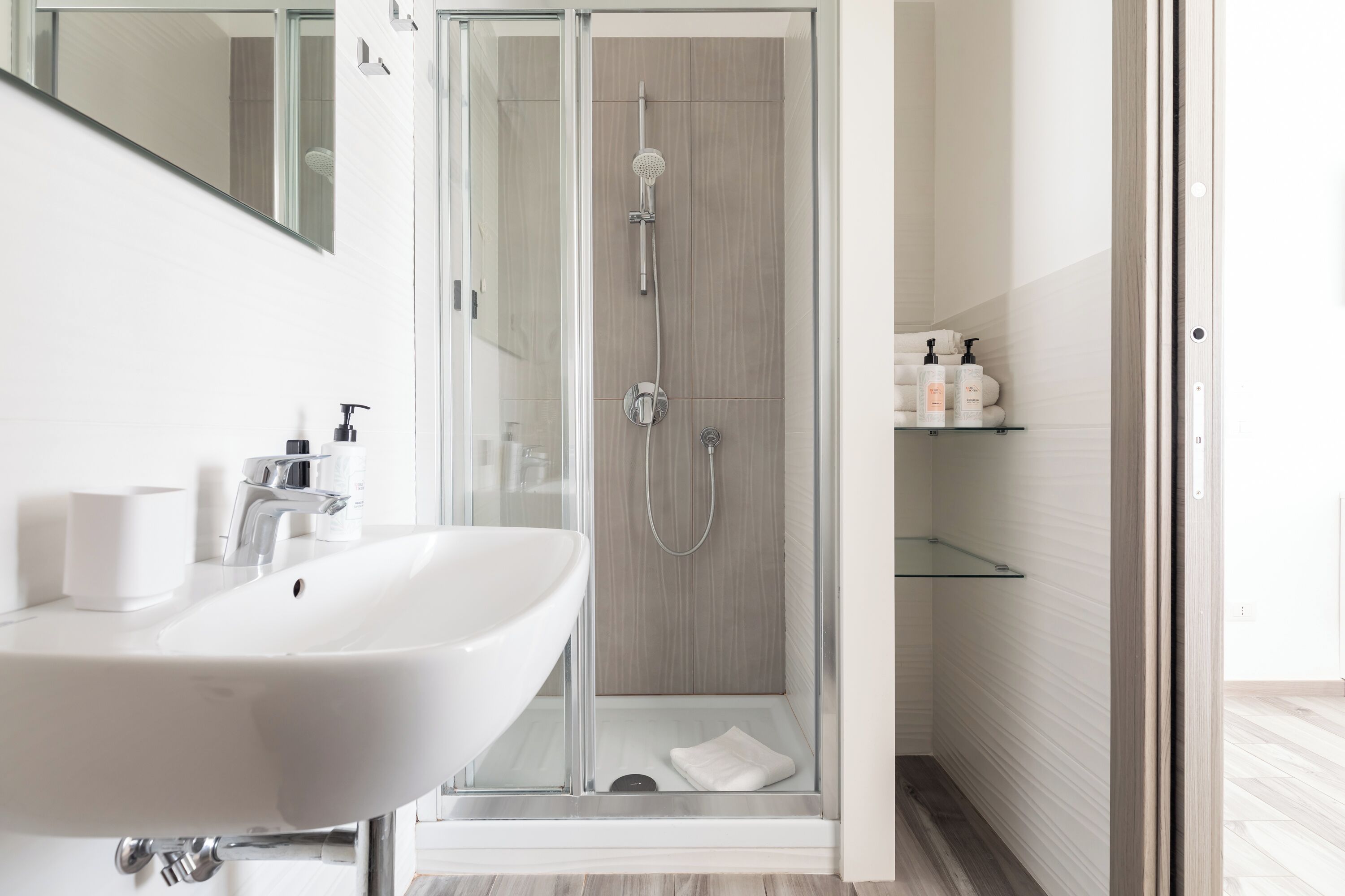 A modern bathroom with a glass-enclosed shower, a white sink, and a glass shelf with toiletries.