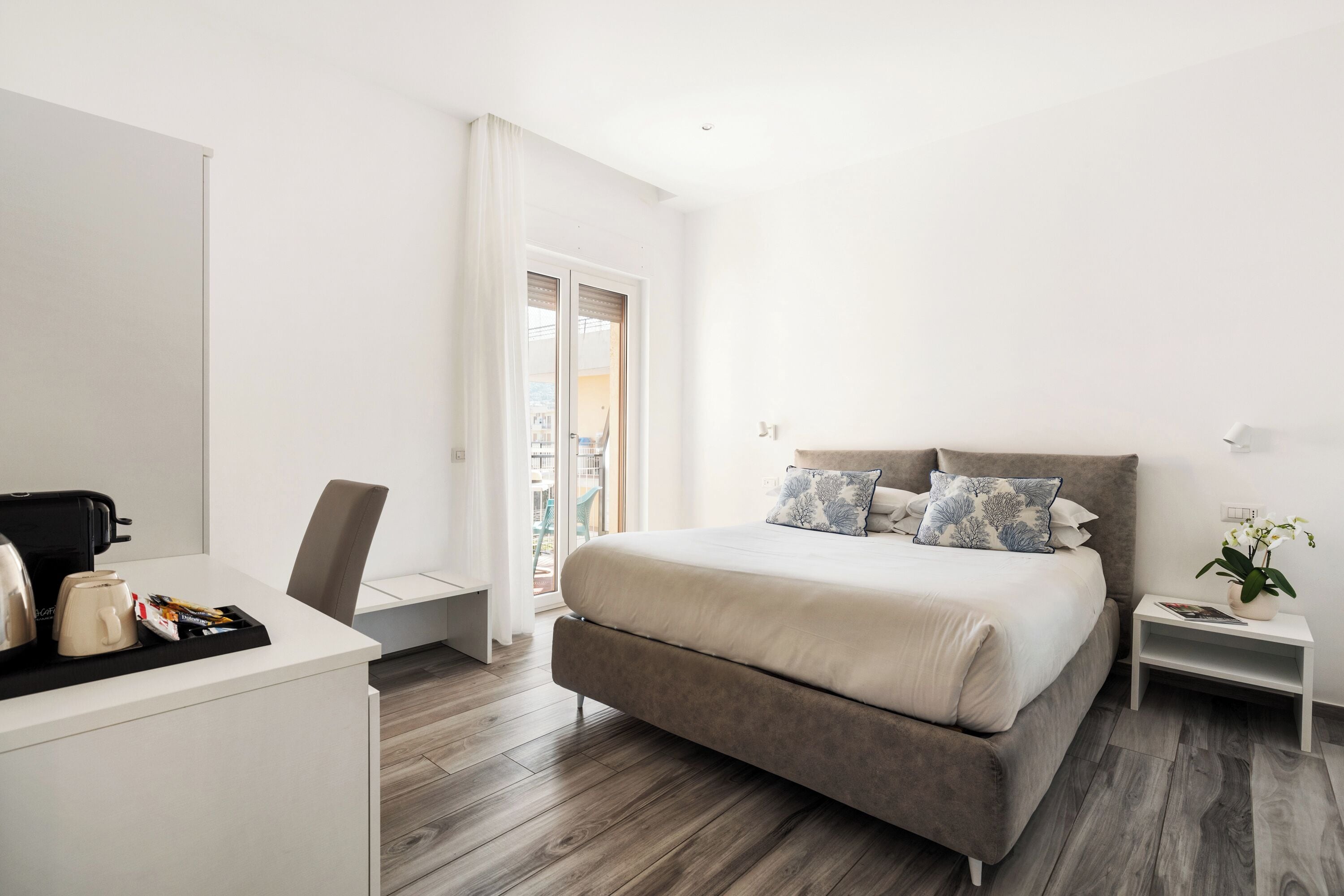 Un dormitorio moderno con una cama grande, un escritorio con cafetera y una mesita de noche con una planta.