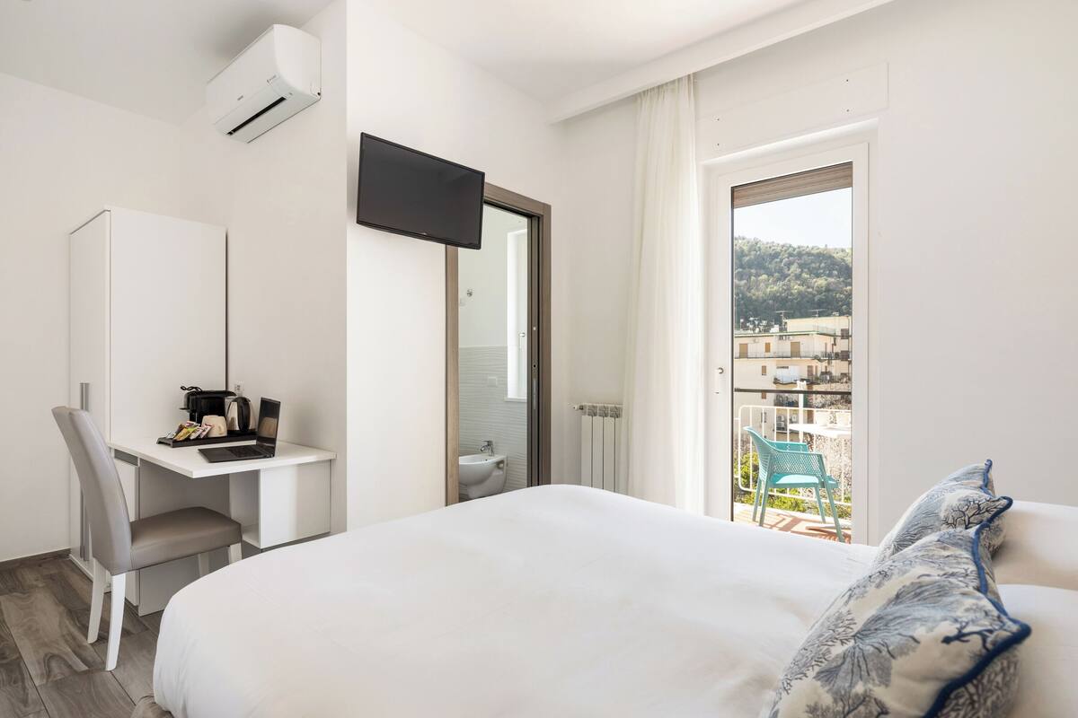 Une chambre d’hôtel comprenant un lit, un bureau avec un ordinateur portable, une chaise, un téléviseur fixé au mur et un balcon offrant une vue sur les bâtiments et les arbres.