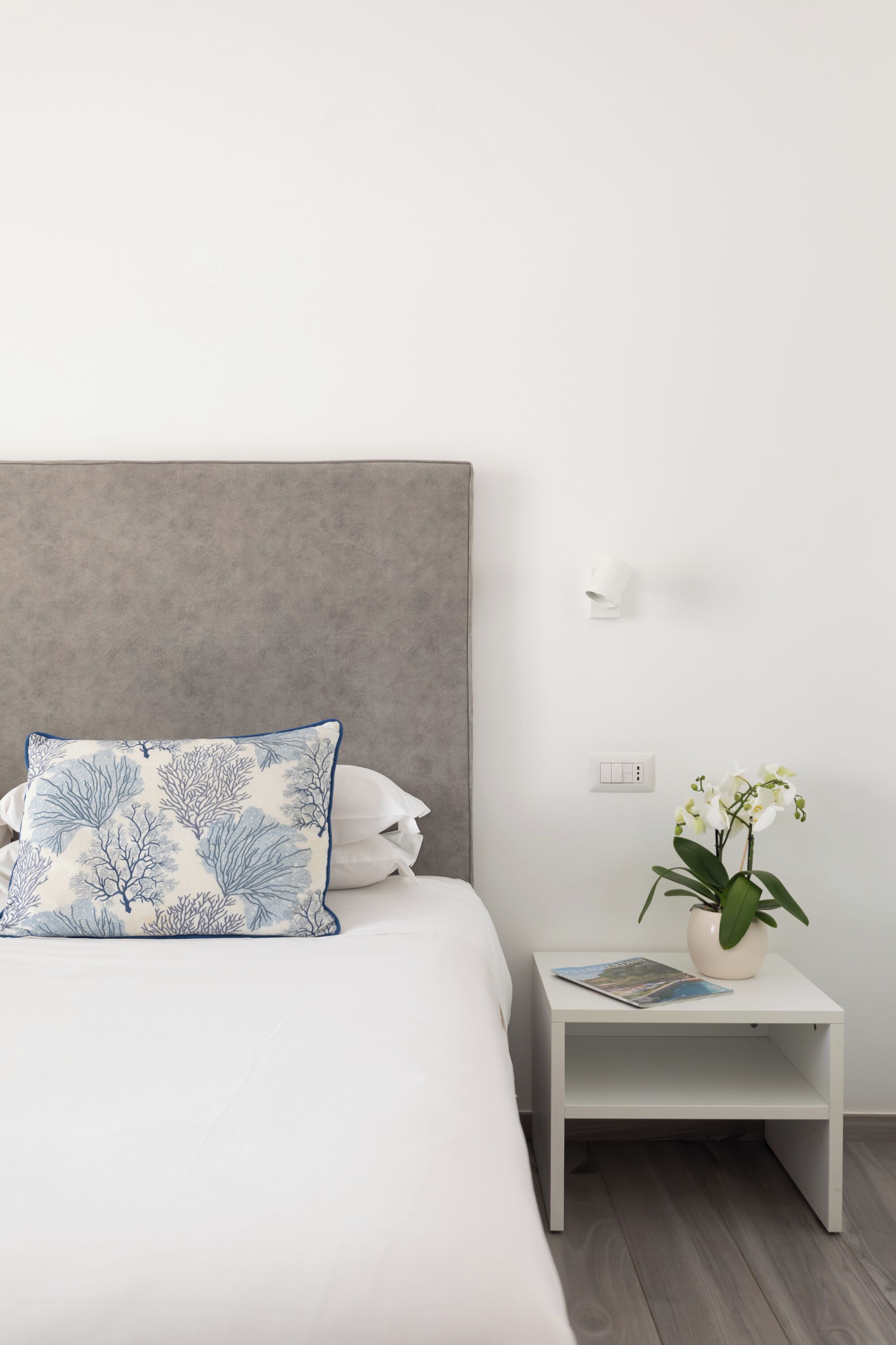 A neatly made bed with a grey headboard, a white nightstand with a plant and a magazine, and a wall-mounted light.