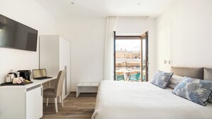 Ein modernes Hotelzimmer mit einem Bett, einem Schreibtisch mit einem Laptop, einem Sessel und Blick auf die Stadt durch ein großes Fenster.