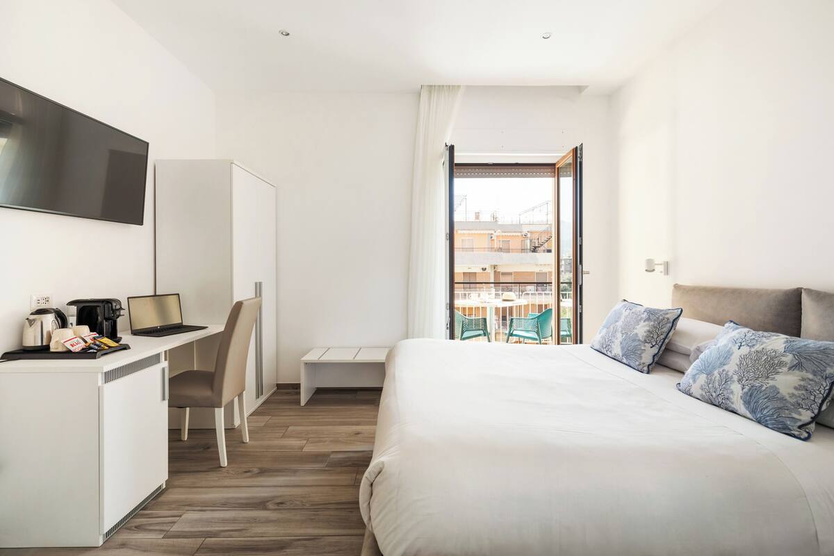 Une chambre d’hôtel moderne, équipée d’un lit, d’un bureau avec un ordinateur portable, d’une chaise et offrant une vue sur la ville grâce à une grande fenêtre.