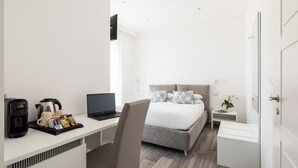 Ein modernes Hotelzimmer mit einem Bett, einem Schreibtisch mit einem Laptop, einer Kaffeemaschine und einem kleinen Nachttisch mit einer Pflanze.
