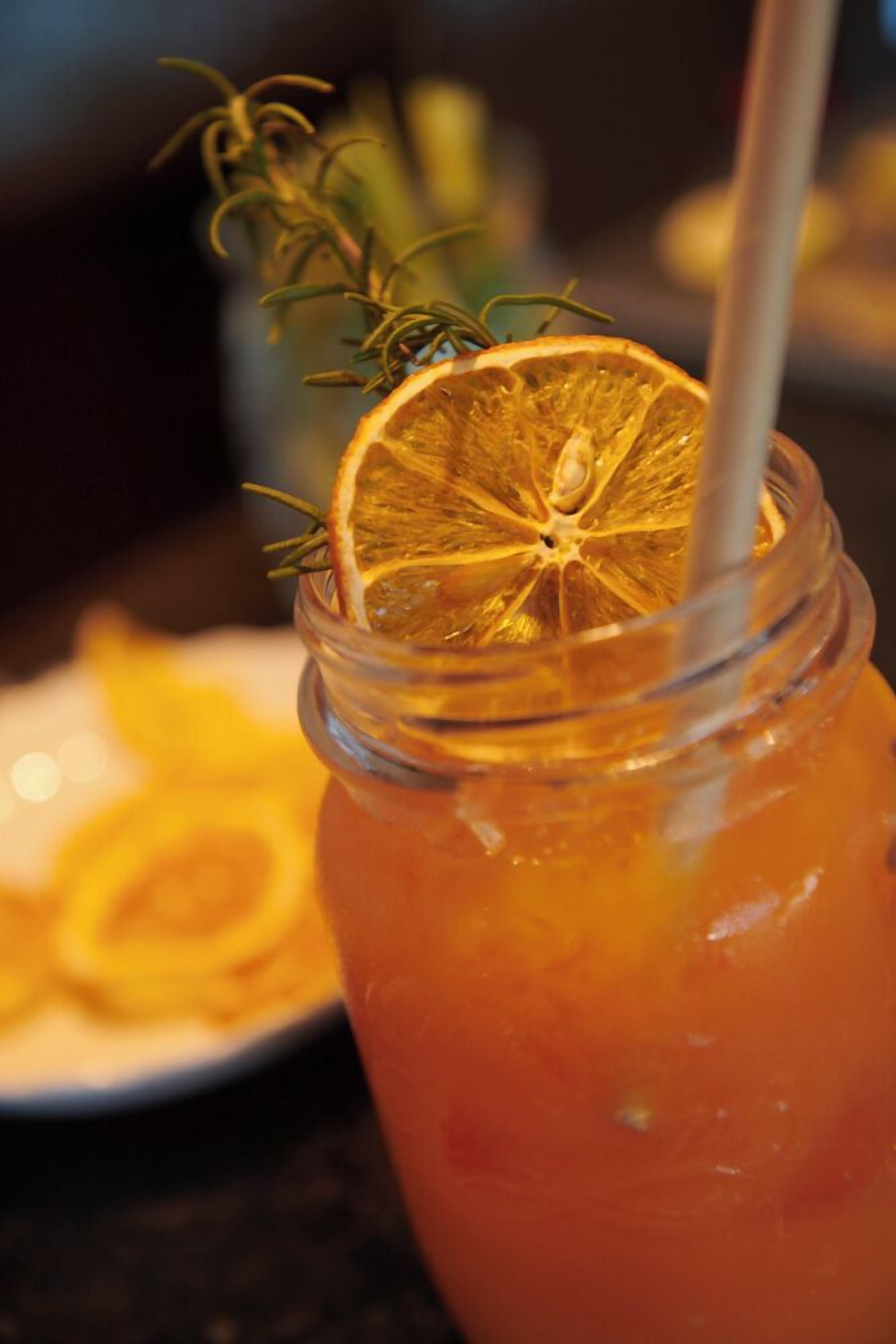 A glass with a drink garnished with a slice of orange and a sprig of rosemary, with a straw.