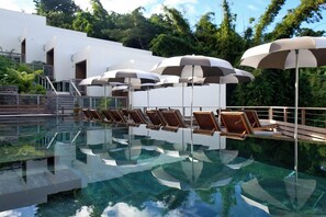Área de piscina com espreguiçadeiras e guarda-sóis, cercada por um edifício branco e vegetação exuberante.