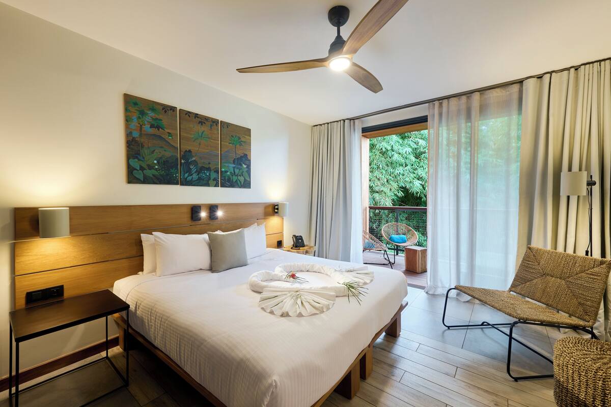 A hotel room with a large bed, a ceiling fan, a balcony with a chair, and artwork on the wall.