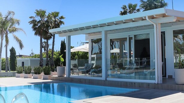 A modern pool area with a glass-enclosed lounge, palm trees, and a clear blue pool. - Female Resort (San Felice Circeo)