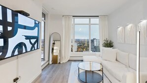 Een moderne woonkamer met een groot raam, een witte bank, een glazen salontafel en een aan de muur bevestigde televisie.