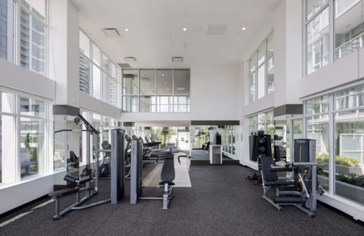 A modern gym with various exercise machines, large windows, and a spacious layout.