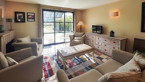 Ein Wohnzimmer mit einer Sofagarnitur, einem Couchtisch, einem Fernseher und einem Schrank. Ein Fenster mit Vorhängen bietet Blick ins Freie.