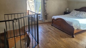 A bedroom with a wooden bed, a nightstand, a lamp, a window with curtains, and a wooden staircase.
