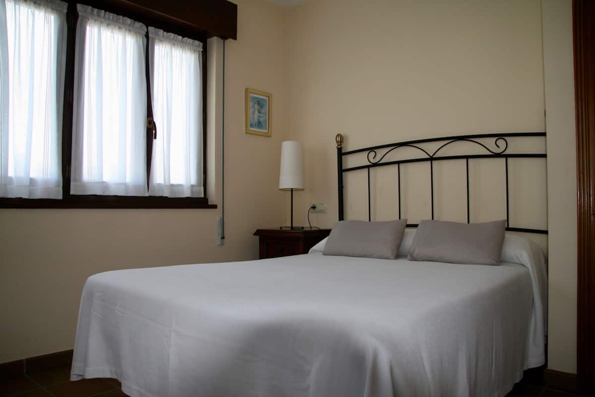 A neatly made bed with a white bedspread and two pillows, a bedside table with a lamp, and a window with curtains.