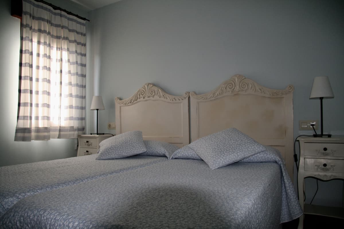 A neatly made bed with a patterned bedspread, two pillows, a headboard with decorative carvings, bedside tables with lamps, and a window with striped curtains.
