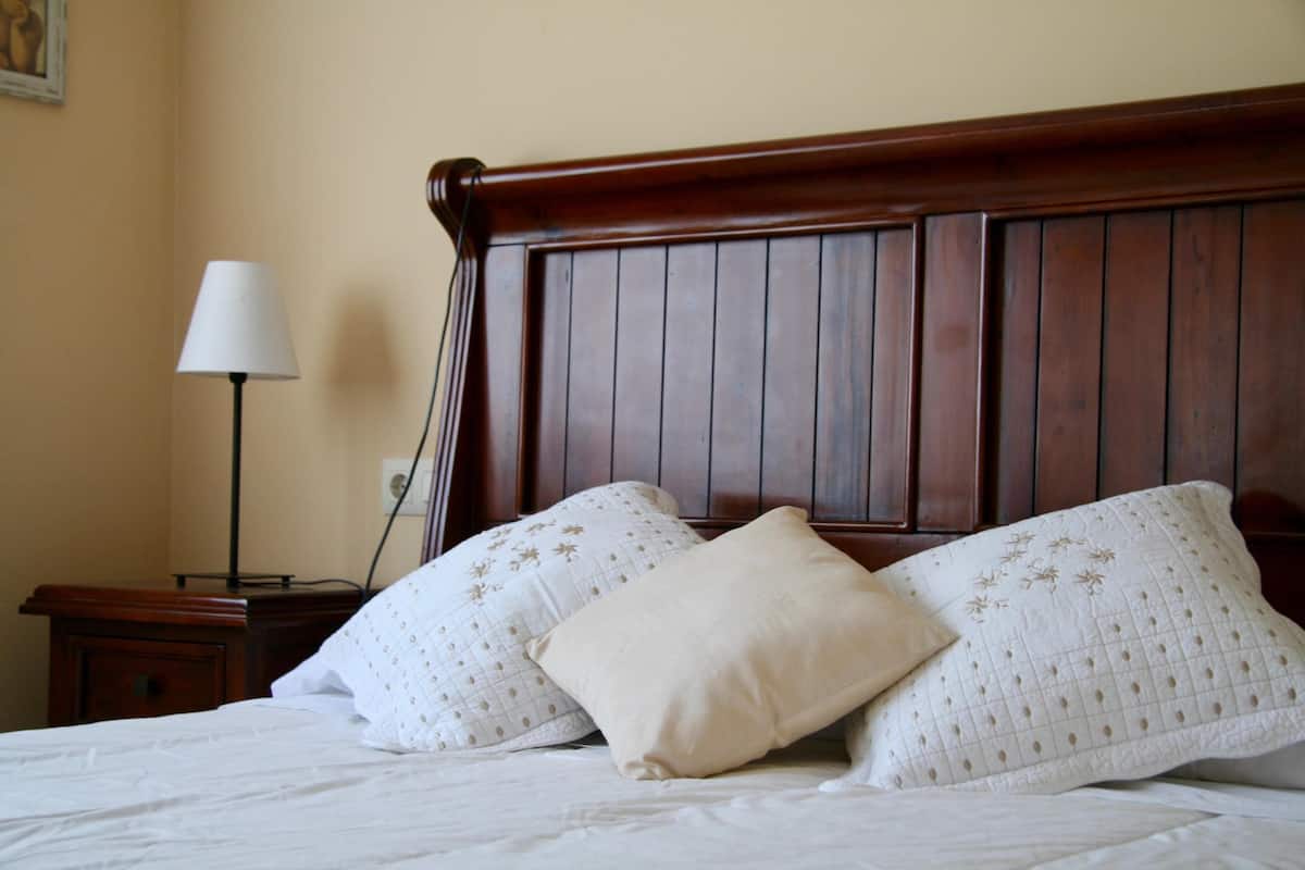 A bed with two pillows, a wooden headboard, a nightstand with a lamp, and a wall with a painting.