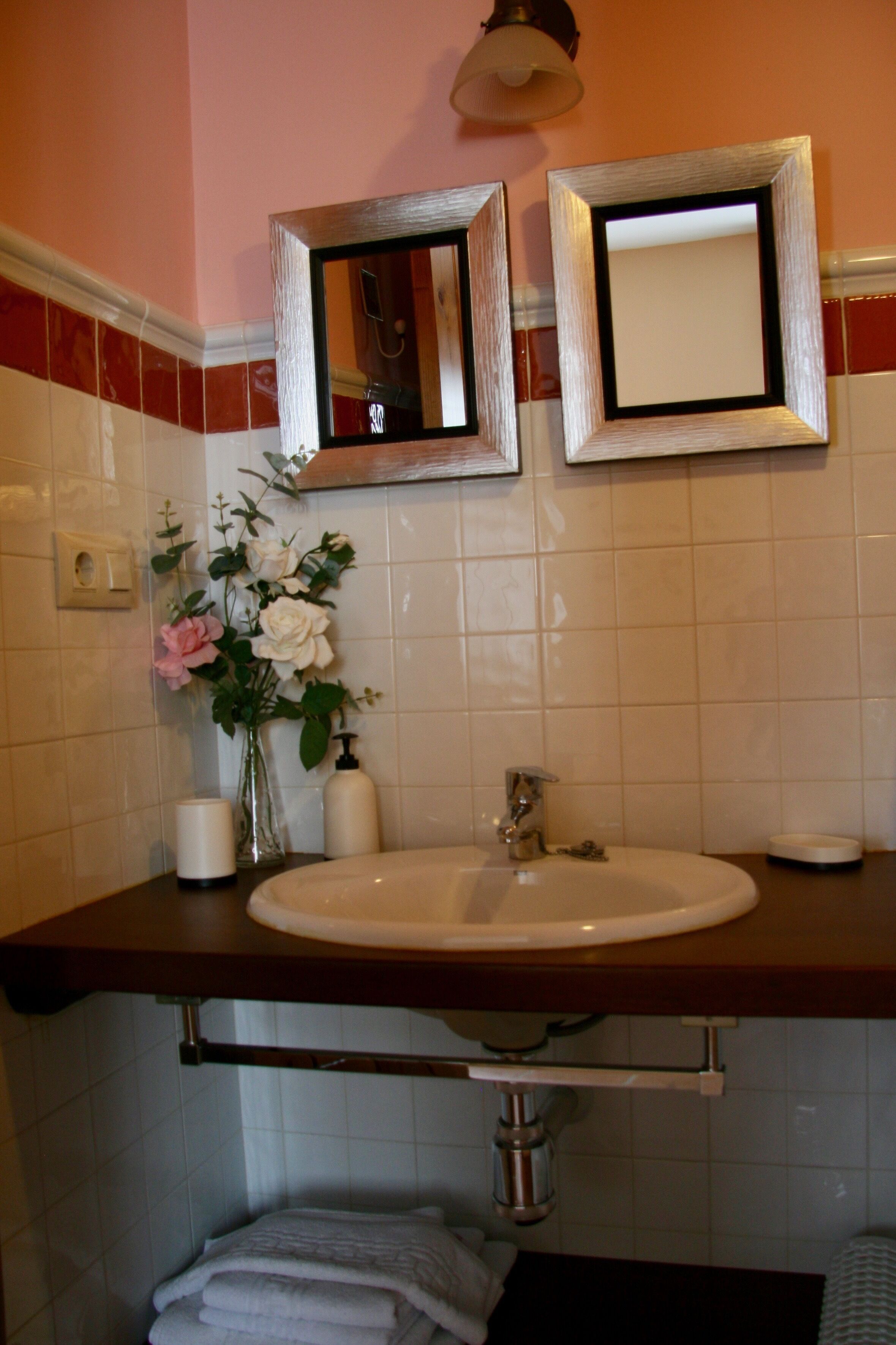 A bathroom with a sink, vanity, and a mirror. There are towels stacked below the sink and a vase with flowers on the vanity.