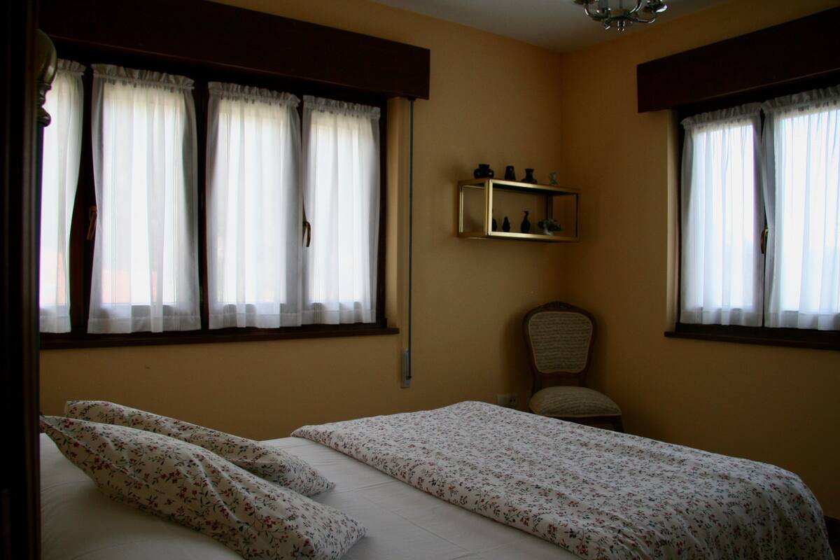 A bedroom with two beds, a chair, a shelf with decorative items, and a window with curtains.