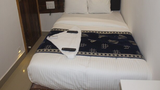 A neatly made bed with white linens and a dark patterned blanket, two remote controls on the bed, and a wooden headboard. - Tanvi Guest House (Mumbai)