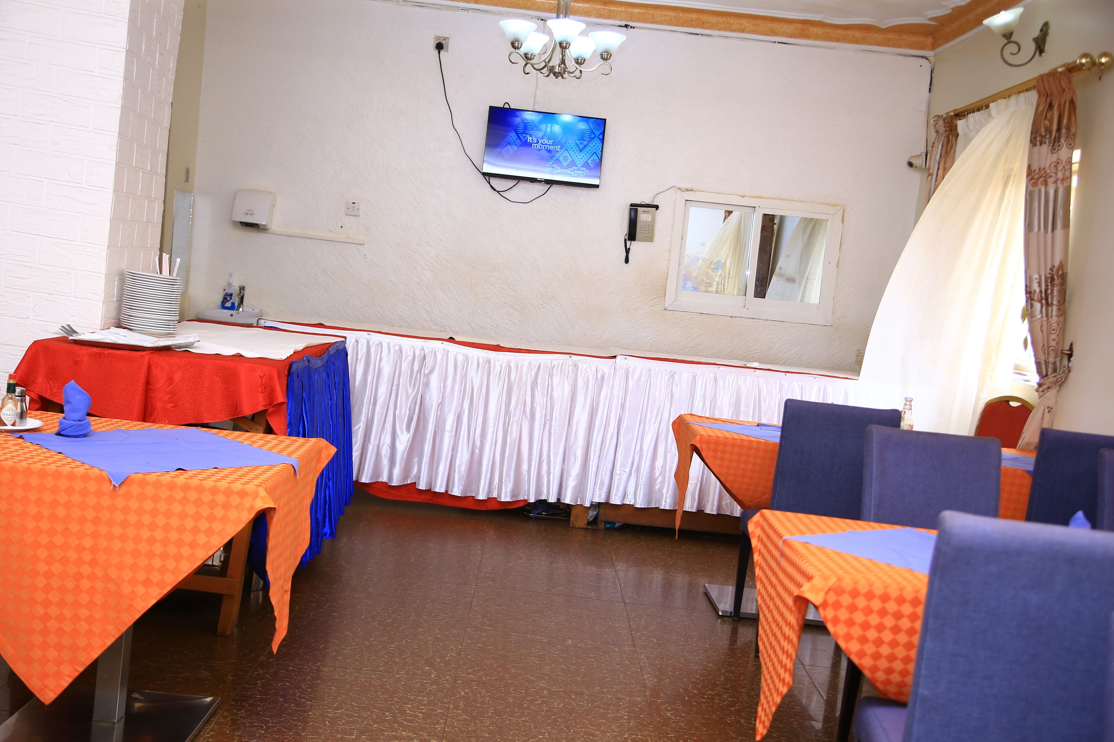 Un espace repas avec des tables recouvertes de nappes à carreaux orange et bleu, un téléviseur fixé au mur et un lustre.