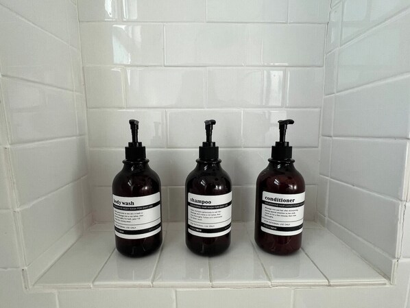 Trois bouteilles de gel douche, de shampoing et d’après-shampoing sont posées sur une étagère carrelée de carreaux blancs.