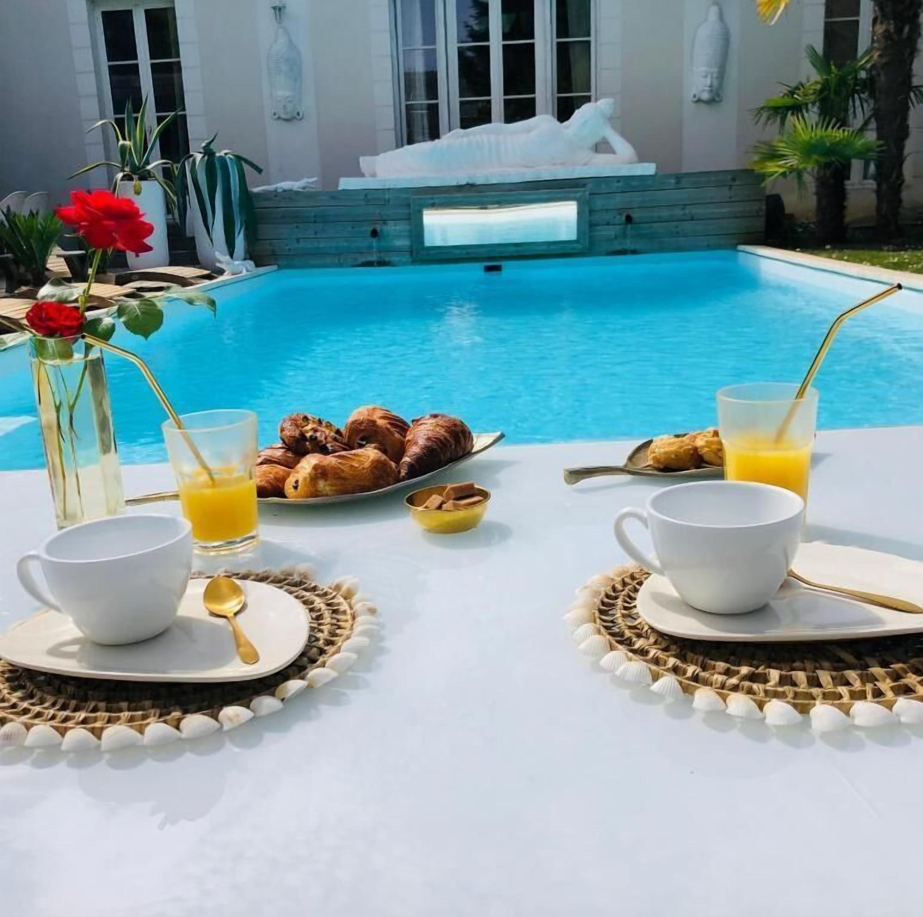 En frukostservering vid poolen med croissanter, apelsinjuice och kaffekoppar på ett bord.