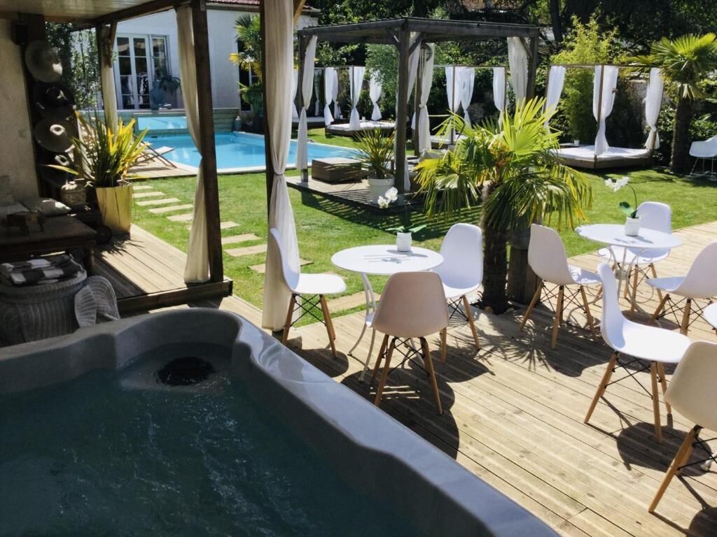Un área de jacuzzi con piscina al fondo, rodeada de sillas blancas y una pérgola con telas colgantes.