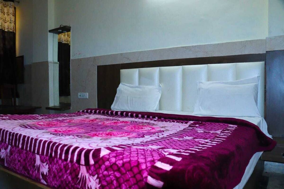 Uma cama com cabeceira branca e almofadas, um cobreleito estampado e uma mesa de cabeceira com abajur.