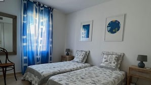Ein Schlafzimmer mit zwei Betten, einem Sessel, einem Nachttisch und einem Fenster mit Vorhängen.