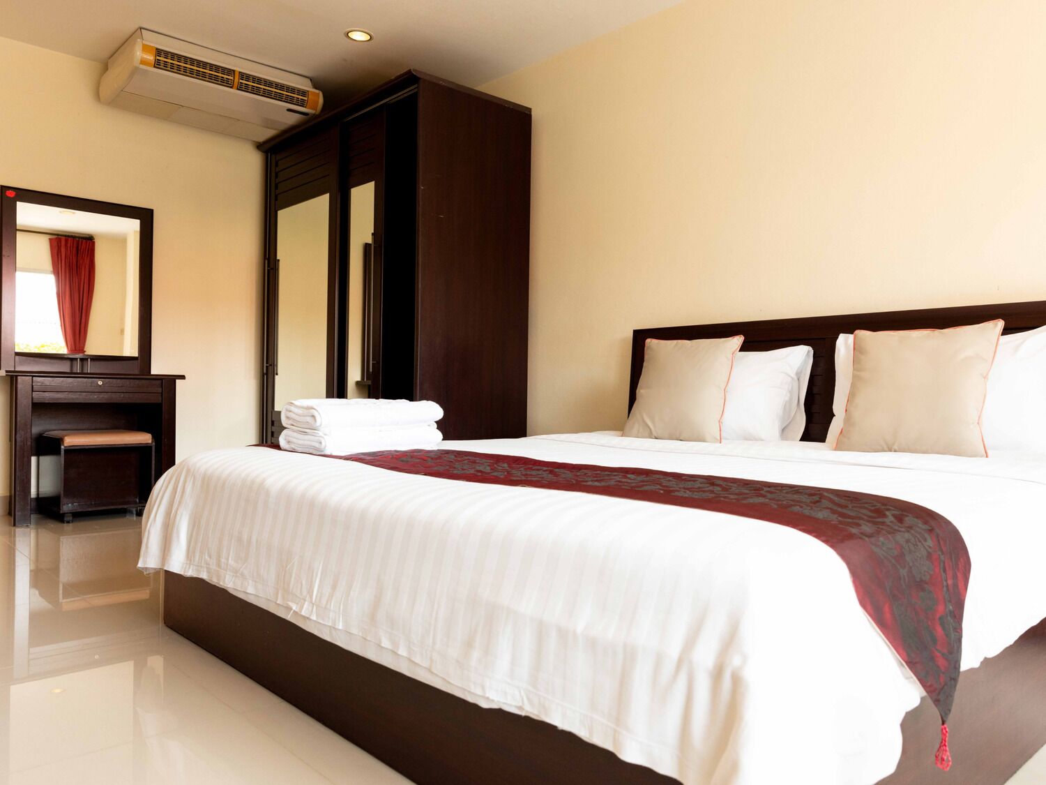 A hotel room with a large bed, a wooden wardrobe, a mirror, and an air conditioning unit.