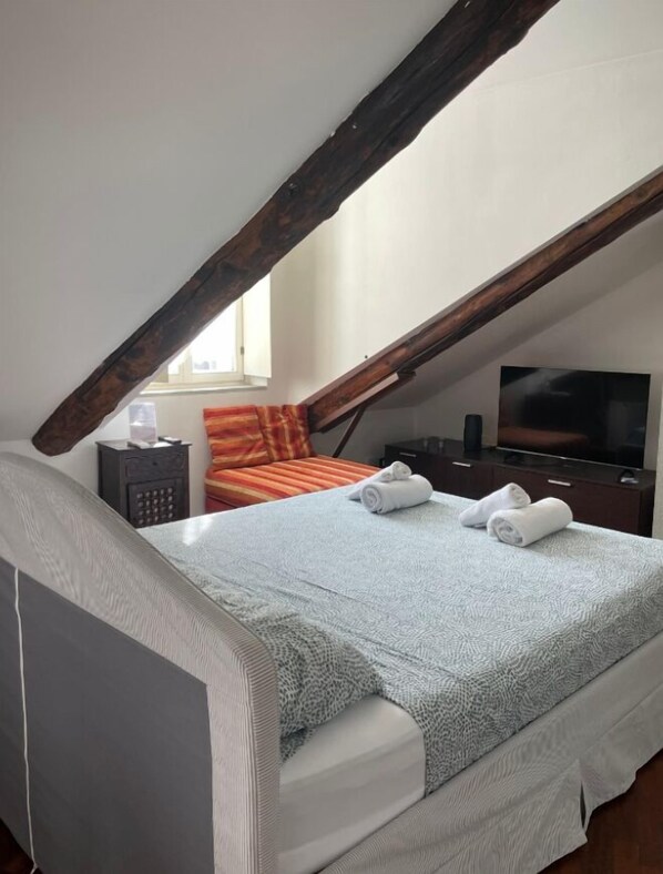 Un dormitorio con techo inclinado, una cama con colcha estampada, un televisor, una mesita de noche y una ventana.