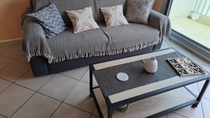 A grey sofa with patterned cushions, a small table with a coaster and a board game, and a view of a balcony through a large window.
