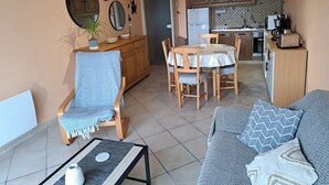 A living room with a sofa, a dining table, and a chair. There is a kitchen in the background with cabinets and a refrigerator.