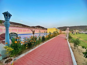A pathway leading to a building with a pool, surrounded by a garden with orange flowers and a clear sky.