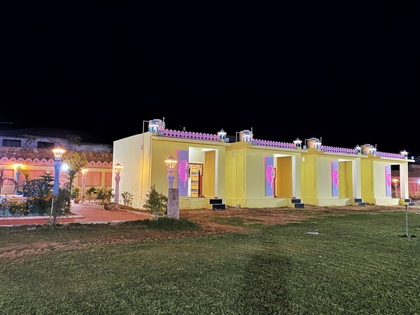 A row of yellow buildings with illuminated entryways and decorative elements, set against a night sky. - The Kukas Resort (Amer)