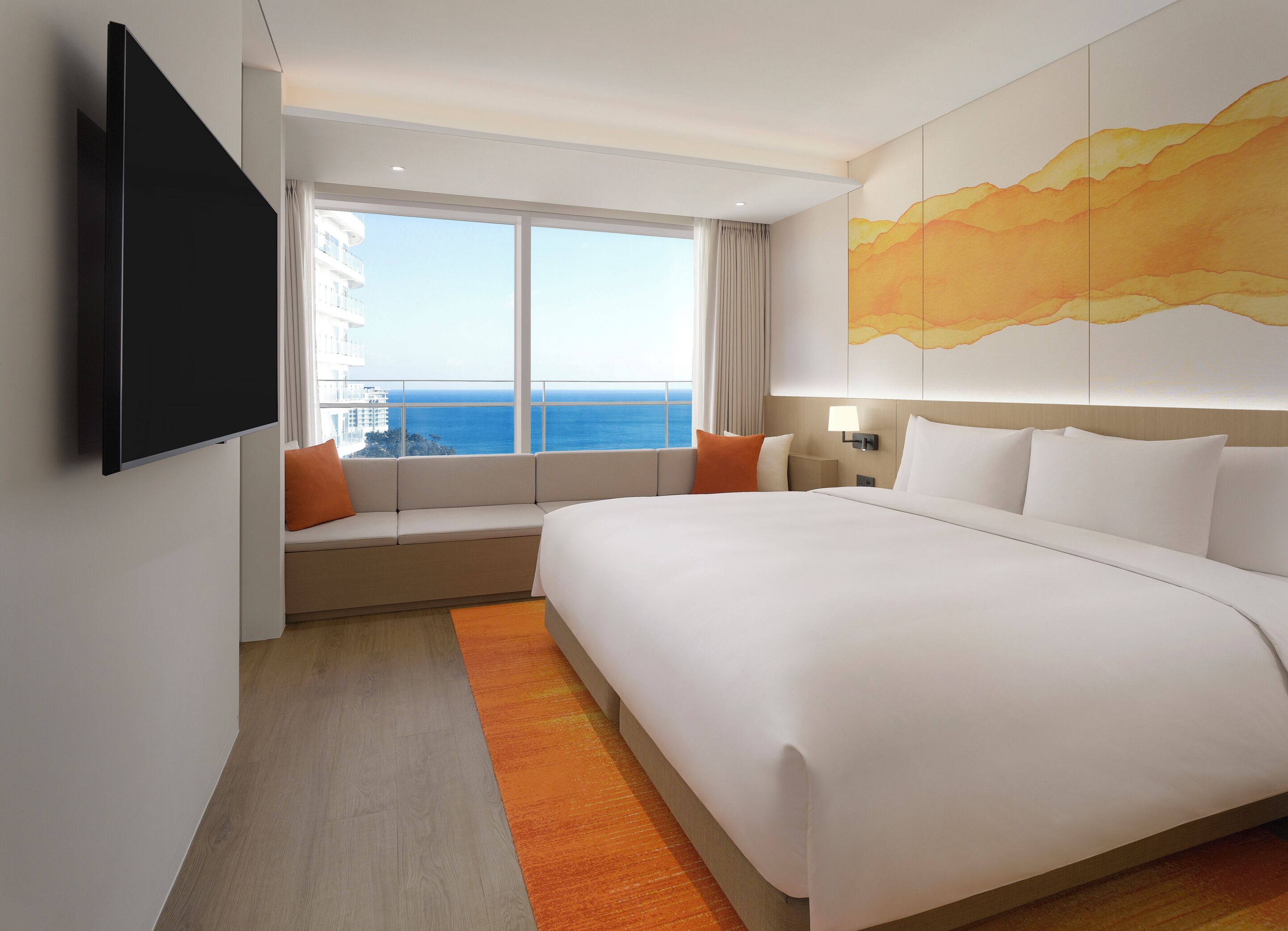 A hotel room with a large bed, a TV mounted on the wall, a window with a view of the ocean, and a painting on the wall.
