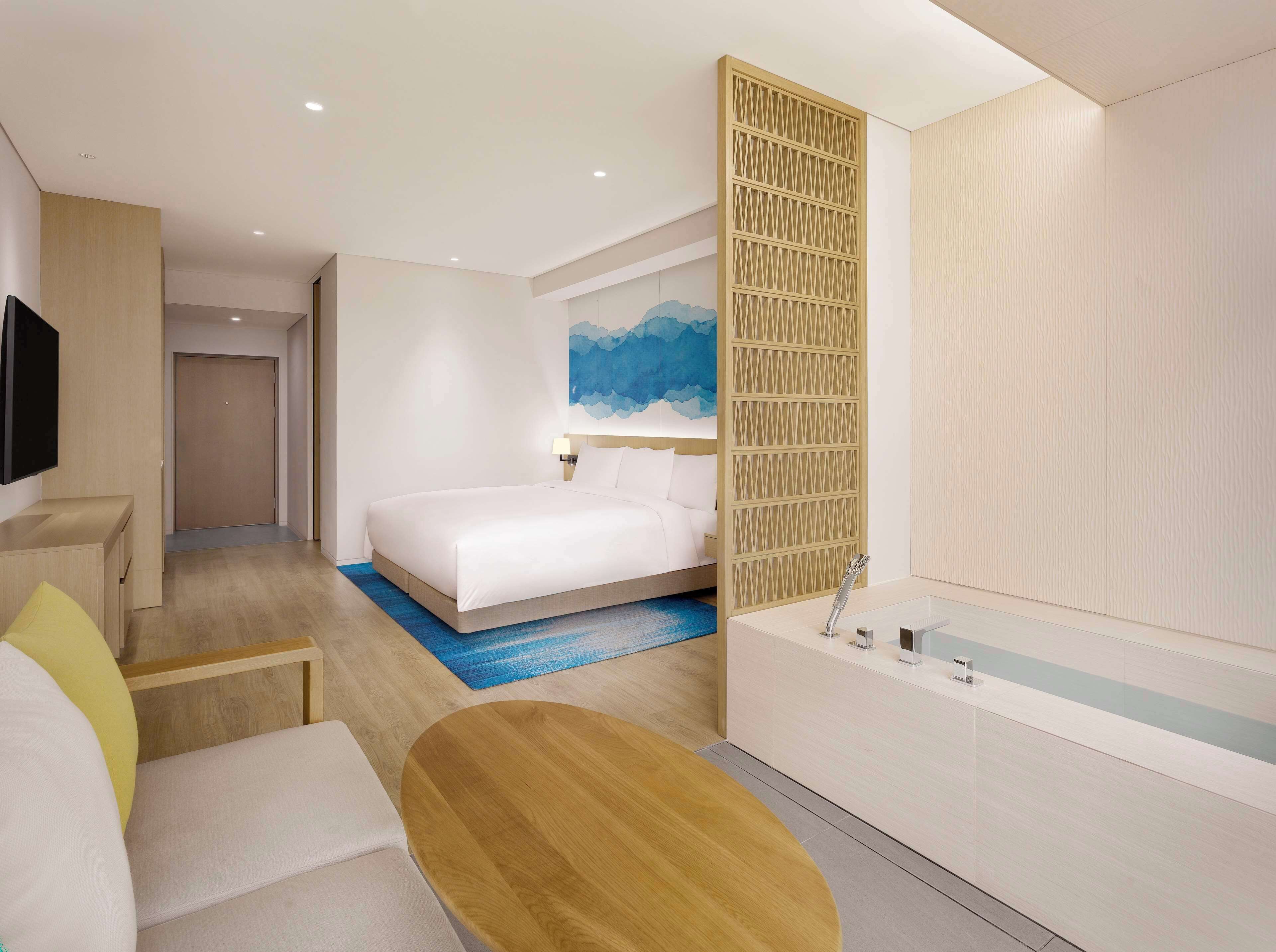 A modern hotel room with a bed, a desk, a chair, a round wooden table, a large window, and a bathroom with a bathtub and sink.