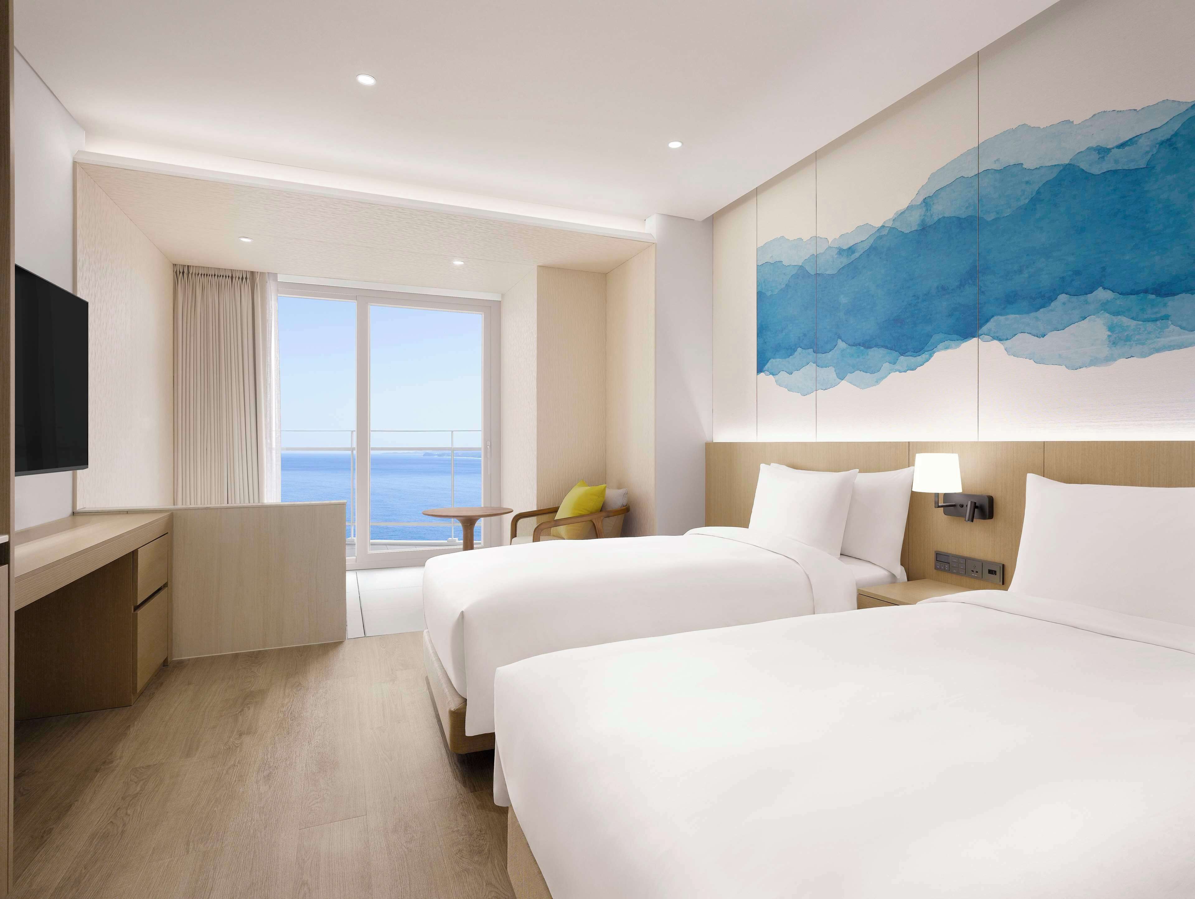 A hotel room with two beds, a desk, a chair, a TV, and a large window with a view of the sea.