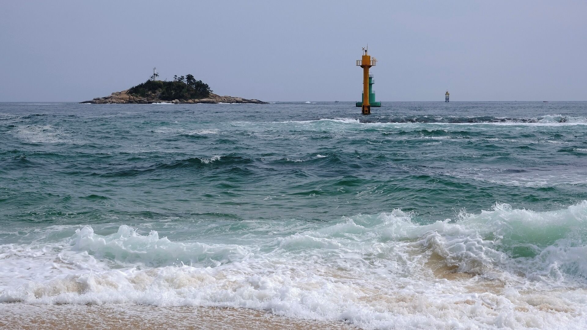A coastal scene with waves crashing onto the shore, a lighthouse, and a small island with vegetation.