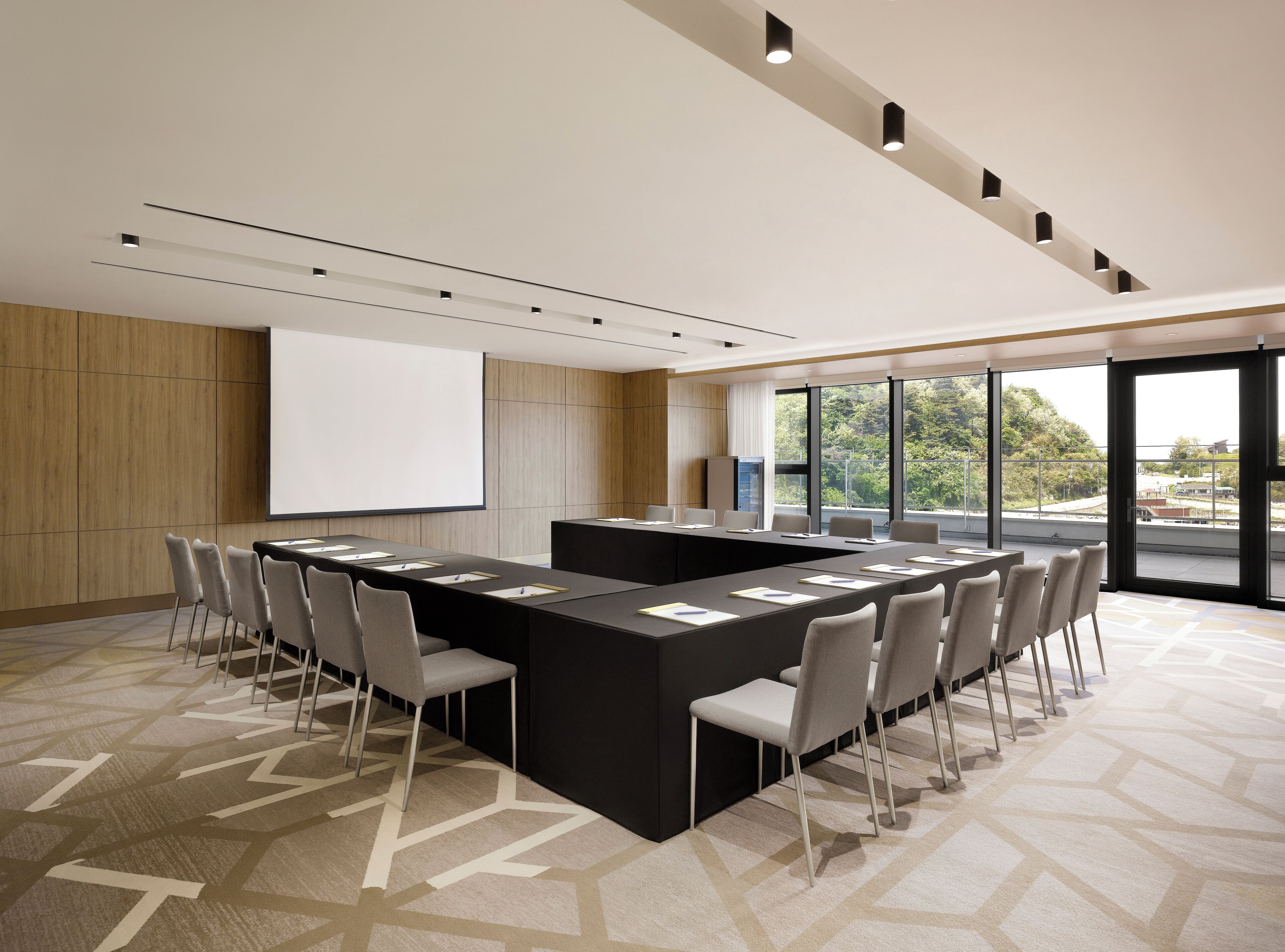 Une salle de conférence équipée d’une grande table, de chaises, d’un écran de projection et de grandes fenêtres donnant sur l’extérieur.
