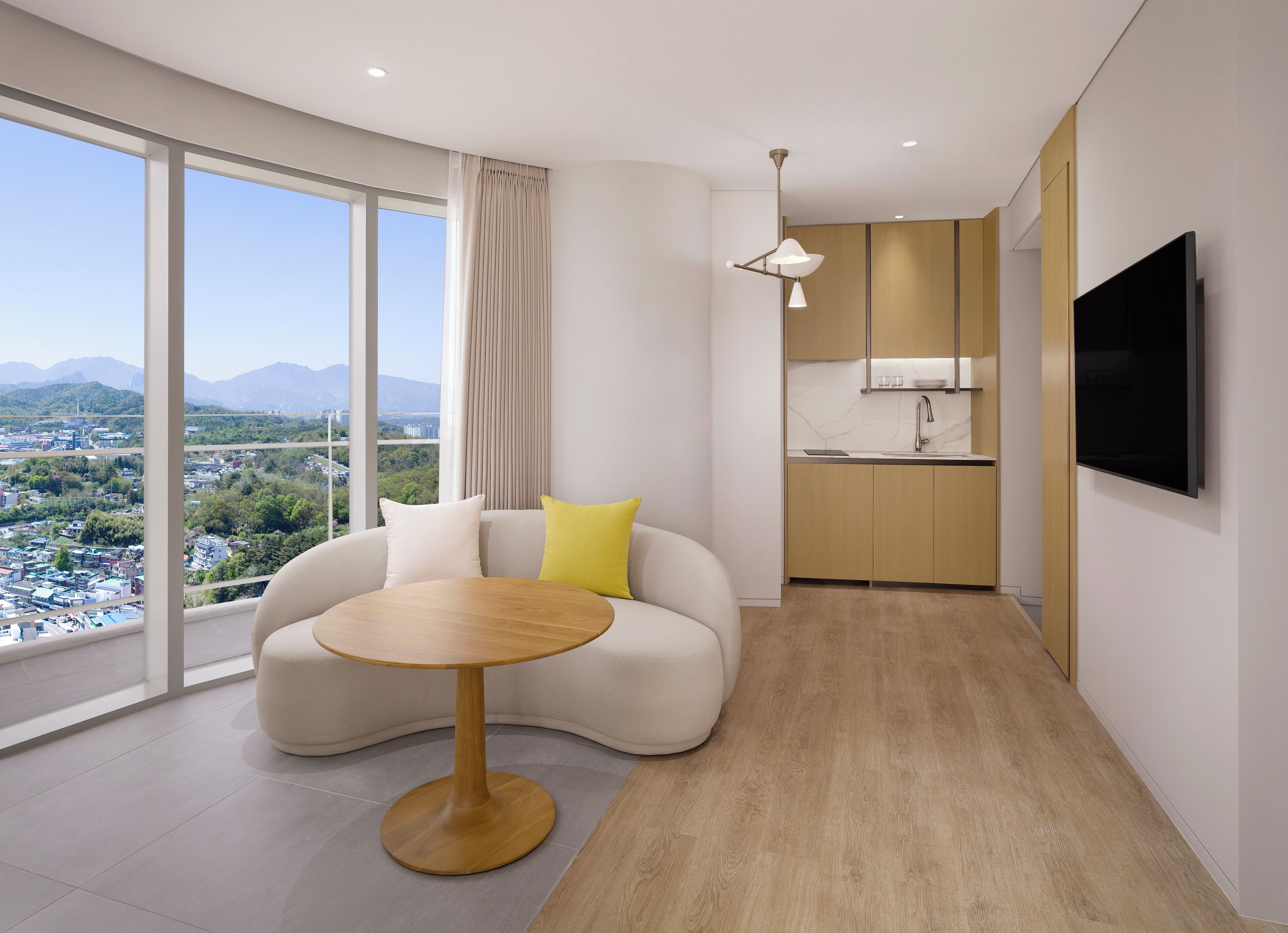 Habitación de hotel moderna con un ventanal grande que ofrece vistas a la ciudad, un rincón de descanso acogedor con una mesa redonda y una kitchenette compacta.