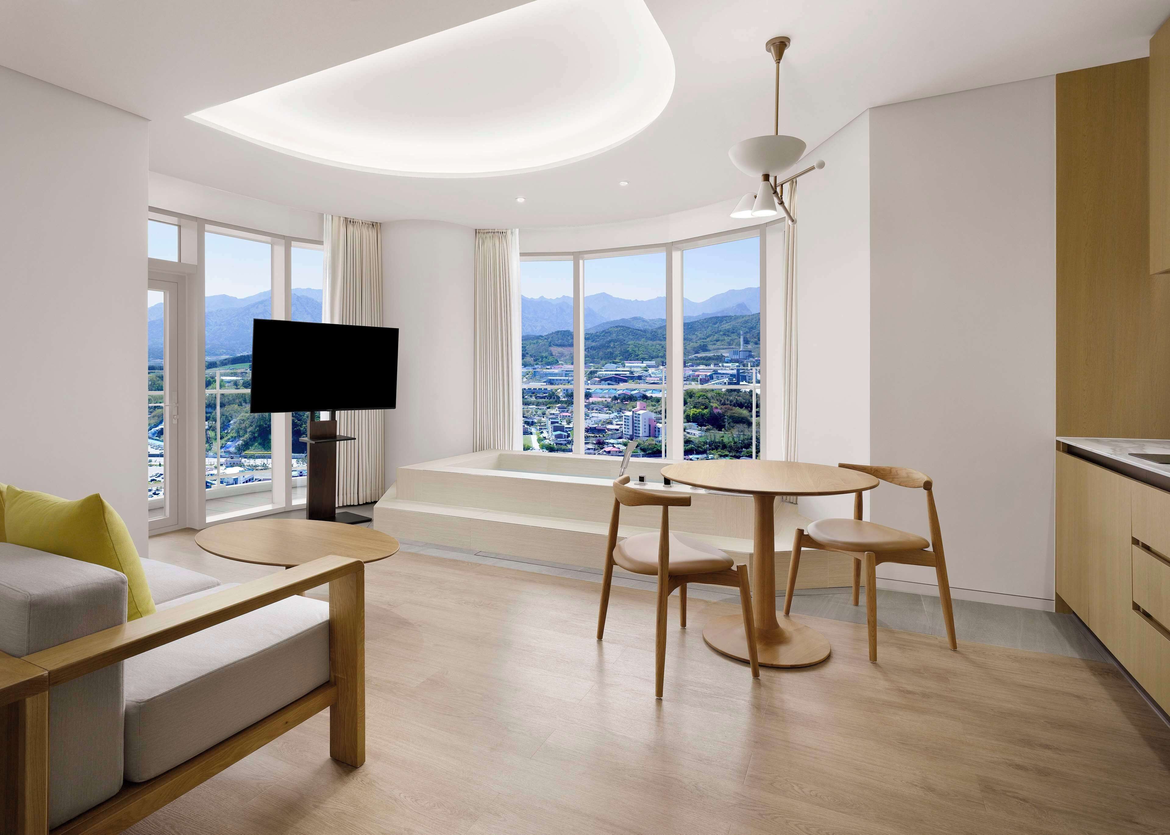 A modern living room with a sofa, a TV, a dining table with chairs, and a view of a cityscape through large windows.