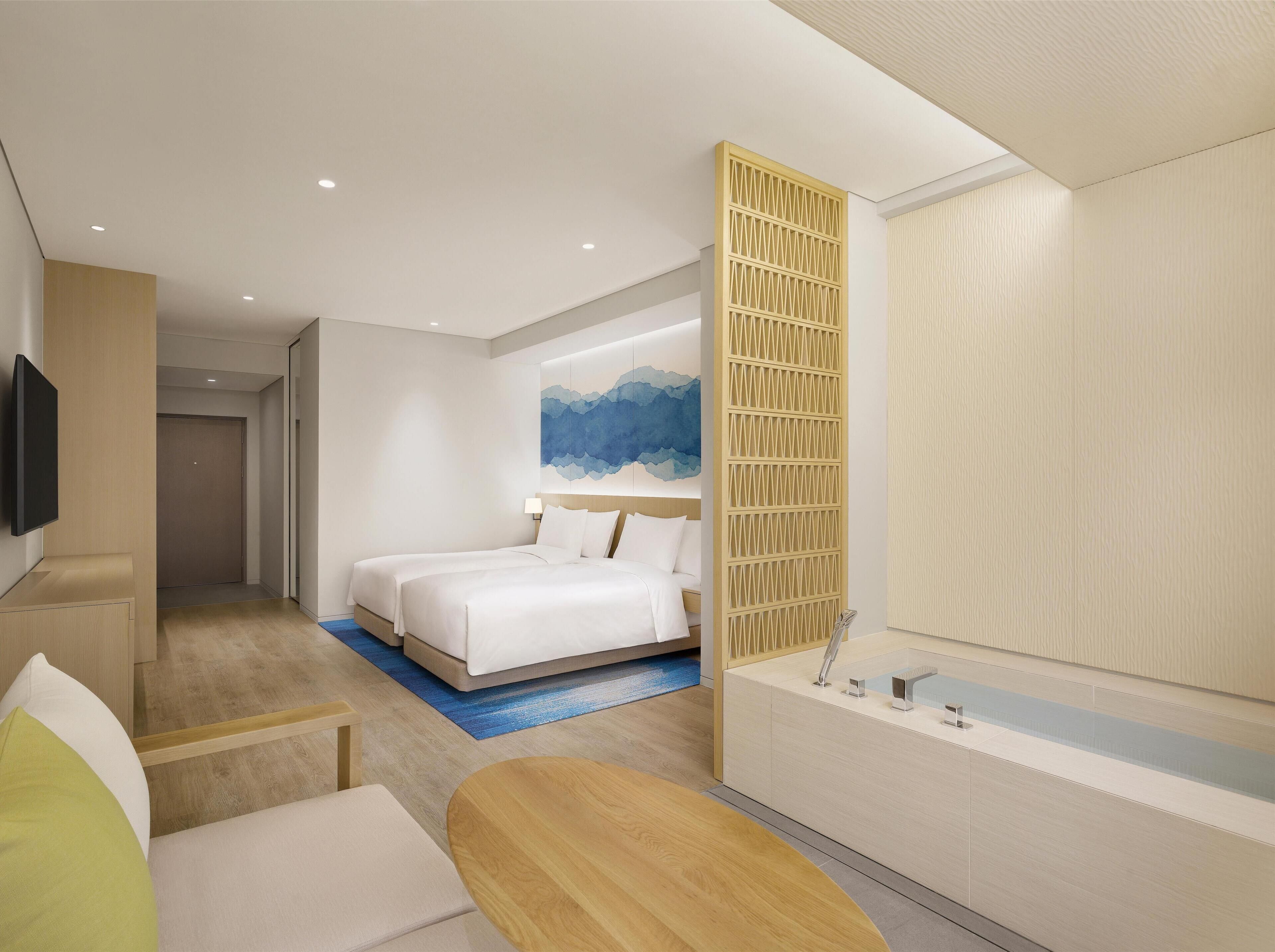 A modern hotel room with a large bed, a wooden side table, a small seating area, and a large window.