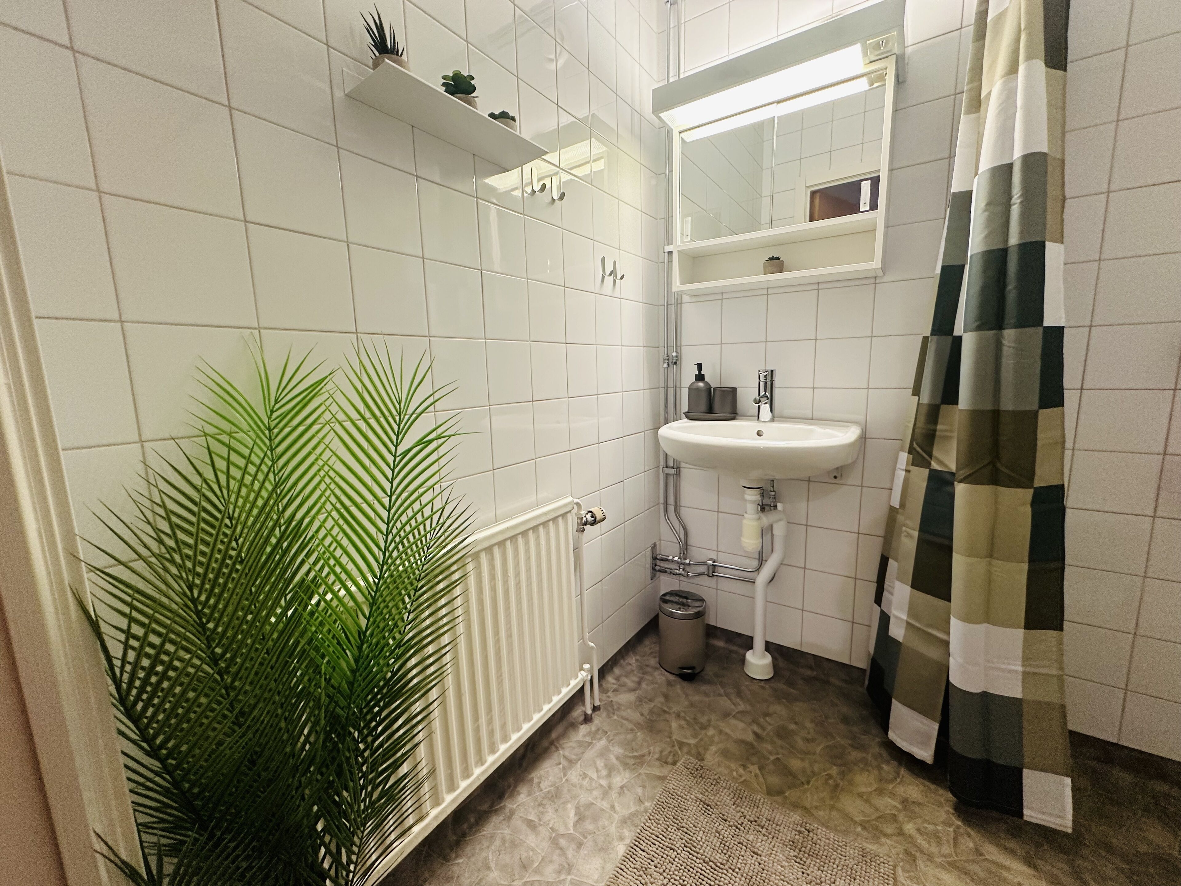 Un baño con azulejos blancos, un lavamanos blanco, un espejo, un radiador, un cubo de basura y una planta verde.