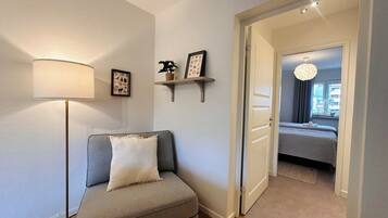 Ett modernt vardagsrum med en grå soffa, en golvlampa och en hall som leder till ett sovrum med en säng och en kristallkrona.
