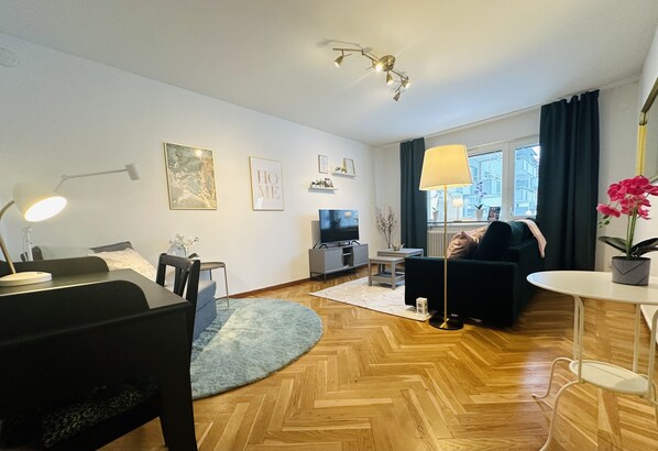 A modern living room with a wooden floor, a black sofa, a dining table, and a TV.