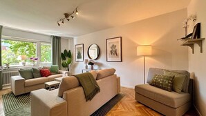 Een moderne woonkamer met twee banken, een salontafel en een vloerlamp. Er is een groot raam met gordijnen, een radiator en een plant.