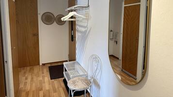Camera d'albergo con una porta in legno, una sedia bianca, un tavolino, uno specchio e una mensola a parete con appendiabiti.
